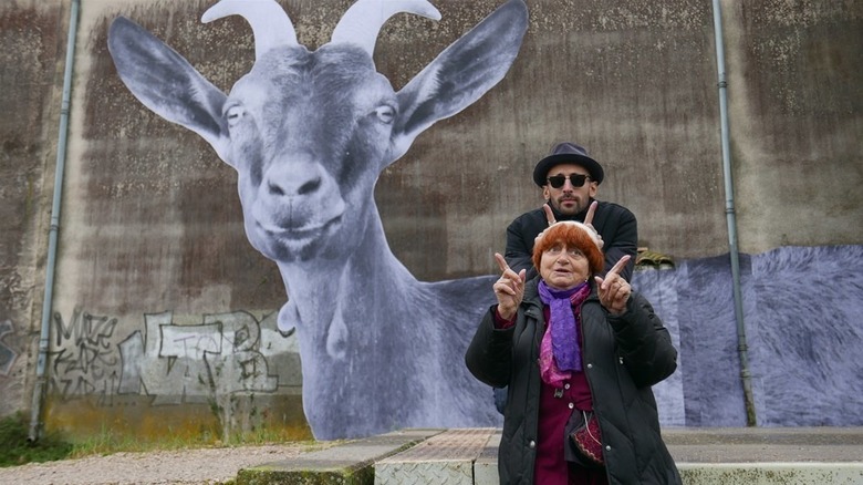 Agnes Varda and JR stand one behind the other, posing with their fingers in an L-shape, in front of a large, outdoor mural of a goat in Faces Places