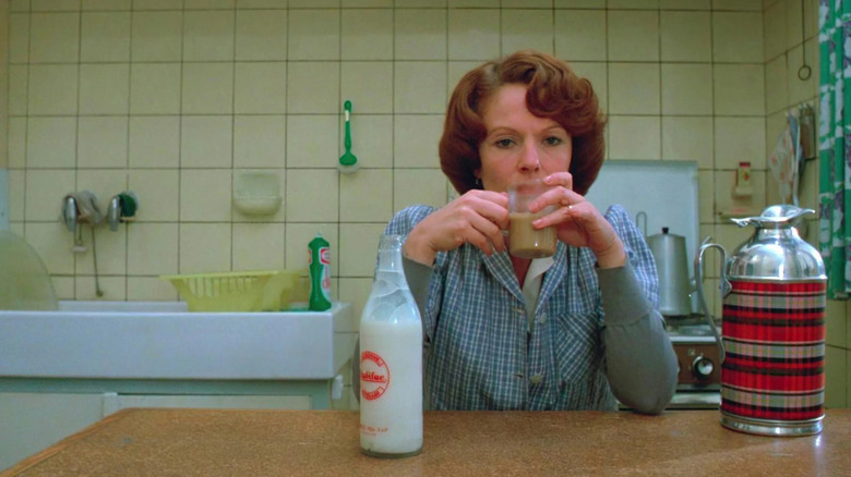 eanne (Delphine Seyrig) sits at the kitchen table with a bored expression, holding a cup in front of her, next to a glass bottle of milk and a plaid-designed thermos in Jeanne Dielman, 23 Quai du Commerce, 1080 Bruxelles