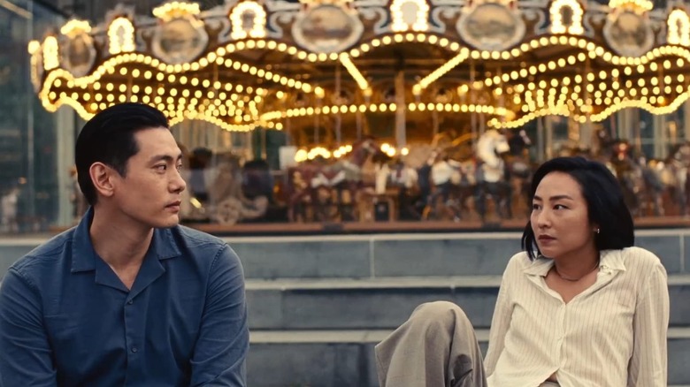 Hae Sung (Teo Yoo) and Na Young (Greta Lee) look seriously at one another while sitting in front of a carousel with glowing lights in Past Lives