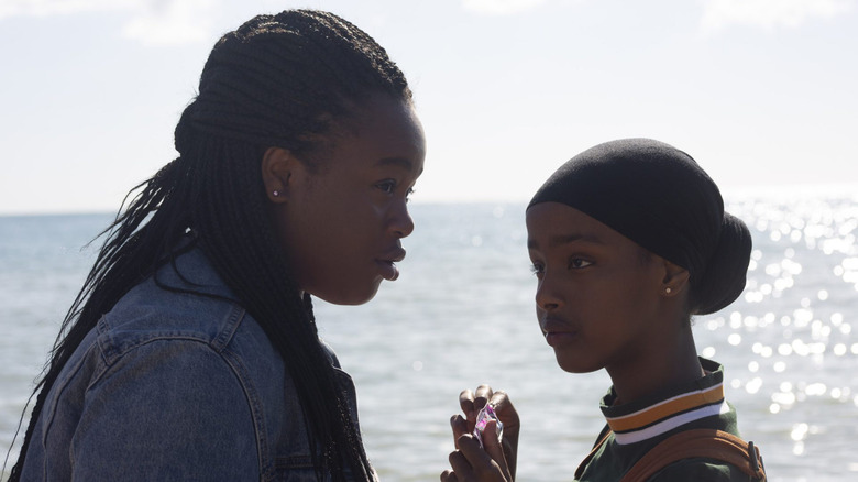 Rocks (Bukky Bakray) looks questioningly into the distance while Khadijah (Tawheda Begum) gazes up at her, with the ocean sparkling in the sun behind them in Rocks