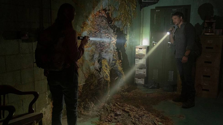 Joel and Tessa point their flashlights at an infected fused to a wall in The Last of Us