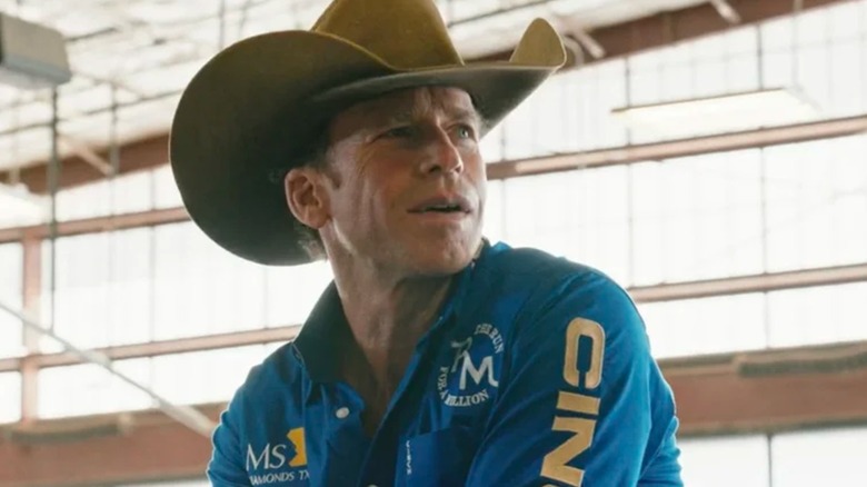 Taylor Sheridan's Travis, in a cowboy hat and branded gear, looks offscreen in Yellowstone