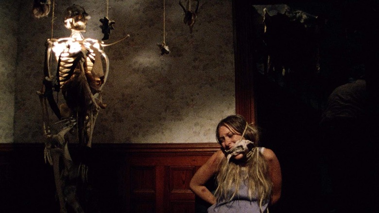 Sally, tied to a chair and gagged, screams next to a lamp made from a human skeleton.