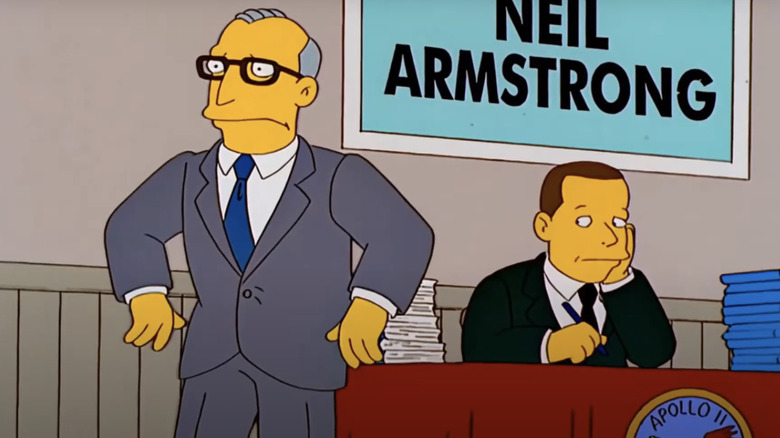 Neil Armstrong sits at a convention desk while his agent stands in the foreground looking upset in The Simpsons