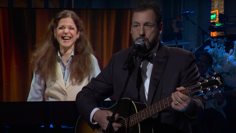 Adam Sandler singing and playing guitar in front of a video of Gilda Radner during SNL 50