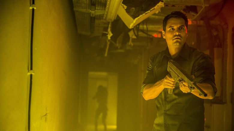 Michael Peña as Peter holding a gun standing in a yellow-lit hallway in Extinction