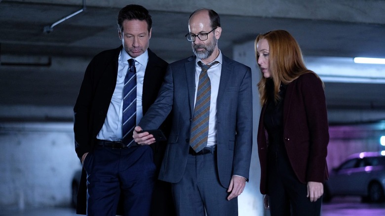 Mulder and Scully looking at the outstretched phone of a bald bearded man standing in between them on The X-Files