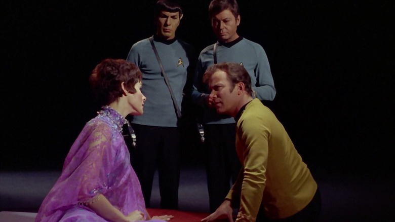 Kirk sits on a platform with a mysterious woman while Spock and Bones look on 