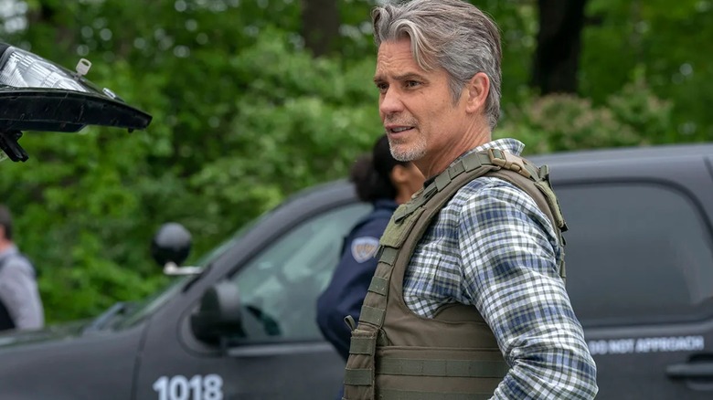 Timothy Olyphant's Raylan Givens, wearing a bulletproof vest, stands beside a police vehicle in Justified: City Primeval