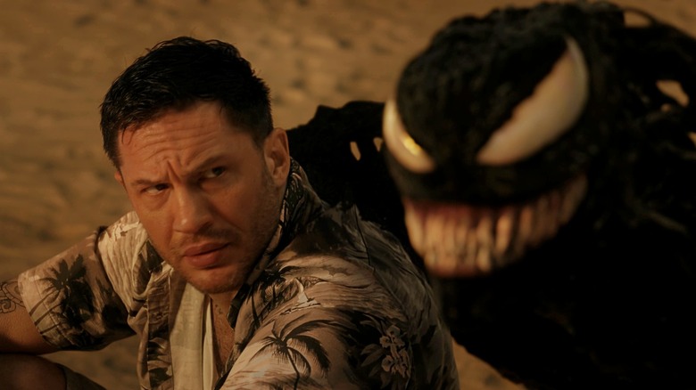 Tom Hardy como Eddie Brock, sentado em uma praia e olhando para Venom consternado