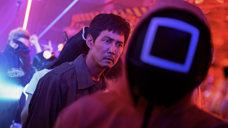 Lee Jung-jae as Seong Gi-hu in a crowded room while he follows a square masked guard in Squid Game season 2