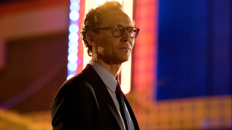 Tom Hiddleston as Chuck, wearing a suit and glasses, in front of colorful lit-up walls, in The Life of Chuck