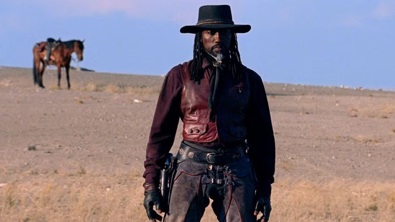 Aman, standing in the desert, a horse behind him in the middle-distance in Gallowwalkers