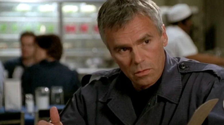 Richard Dean Anderson's Jack sitting at a table with people behind him in Stargate SG-1