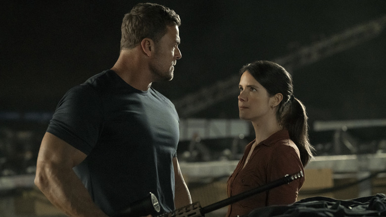 Reacher, wearing a T-shirt, holds a rifle while looking at a brown-haired woman next to him