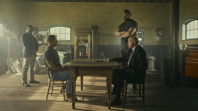 Reacher sits at a table opposite a villain, while a gigantic man looms over them both