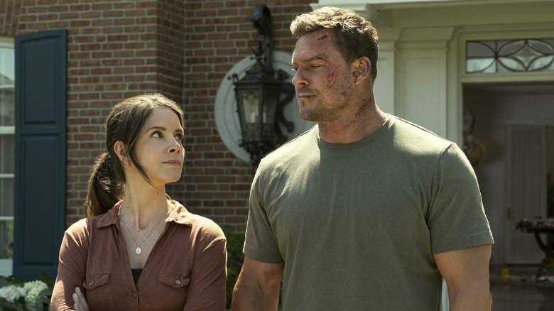 A beat-up Reacher, wearing a grey-green T-shirt, looks at a woman standing beside him