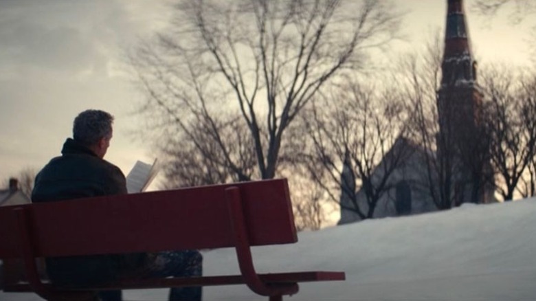 Irving on a park bench in Kier, looking at a book. A church is over the crest of a hill. A shot from Severance