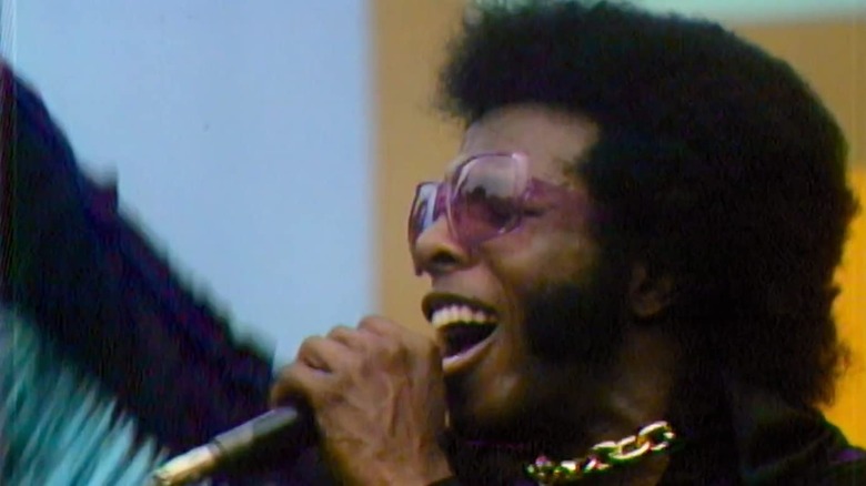 a man with pink sunglasses and a fringe jacket sings into a microphone with his arm raised