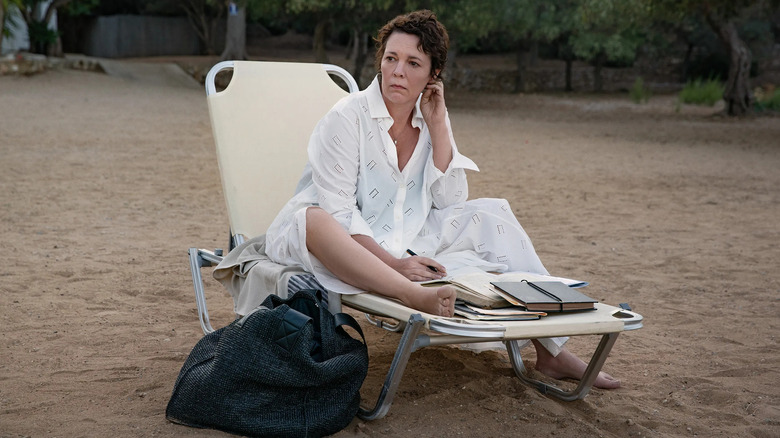 a woman in a white shirt dress sitting on a loung chair on the beach while writing and looking to her right