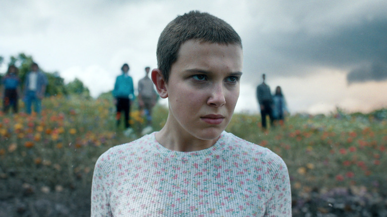 Millie Bobby Brown as Eleven standing in a meadow of flowers and dead grass in Stranger Things season 4