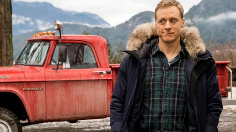 Alan Tudyk's Harry stands in winter wear in front of a dirty red pickup truck in the mountains in Resident Alien