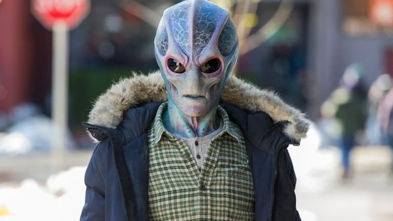 A blue-purple alien wearing a wintery human outfit stands on a street in Resident Alien