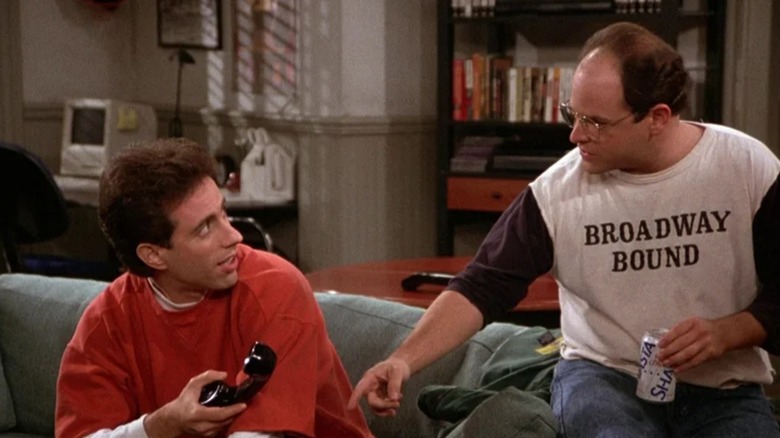 Jerry Seinfeld's Jerry holds up a phone in his apartment while Jason Alexander's George, wearing a shirt that says Broadway Bound, gestures at him in Seinfeld