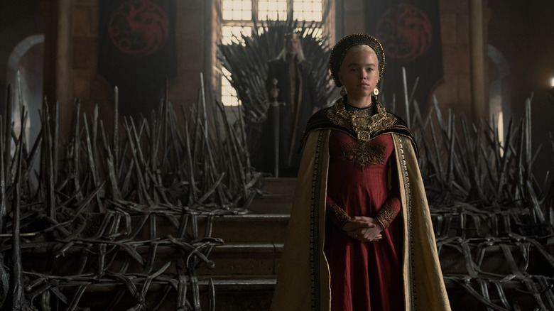 Young Rhaenyra in royal Targaryen garb standing in front of Viserys on the Iron Throne in House of the Dragon