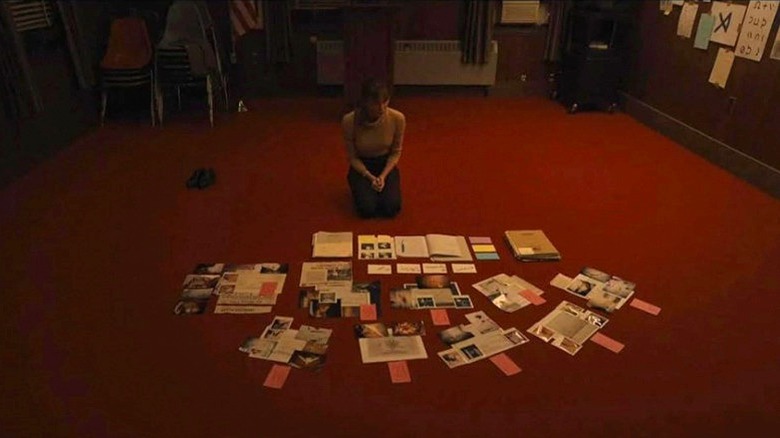 Agent Harker sitting on a red carpet, examining a splayed out series of papers in Longlegs