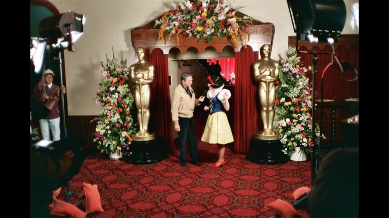 Army Archerd holds a microphone and stands next to an actress dressed as Snow White, standing outside the entrance to the ballroom to the 61st Academy Awards