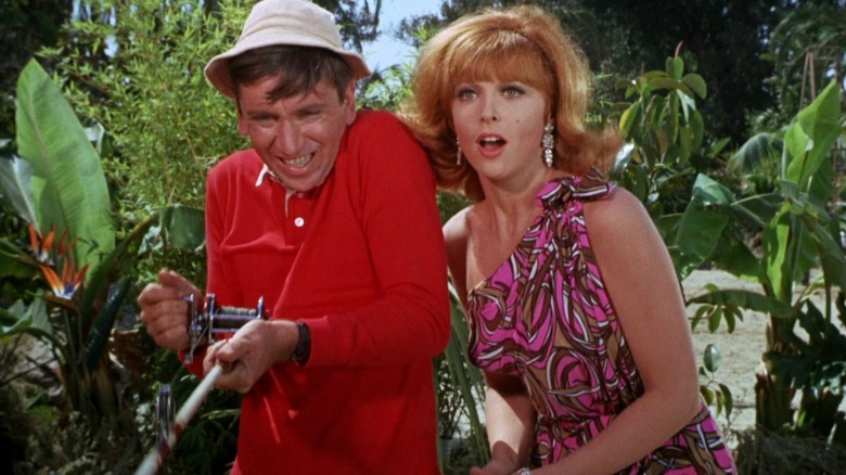 Gilligan striving with a fishing rod, ginger, next to him, encouraging him.