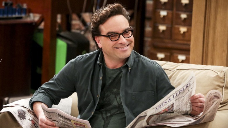 Leonard no sofá lendo jornal e rindo da Teoria do Big Bang