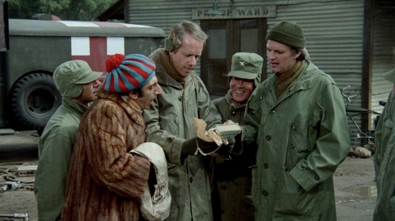 The cast of M*A*S*H gathered around a book wearing cold weather clothes