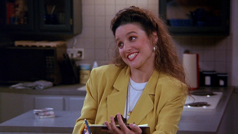 Elaine Benes, smiling wide in a kitchen, holding a clipboard.