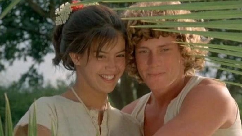 Phoebe Cates' Sarah and Willie Aames' David smiling, looking down at something offscreen through palm frond leaves in Paradise