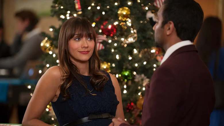 Ann Perkins olha ceticamente para Tom Haverford com uma árvore de Natal iluminada em segundo plano, enquanto eles estão perto de um bar