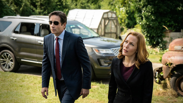 Mulder, wearing sunglasses, walks next to Scully in front of a parked car on The X-Files