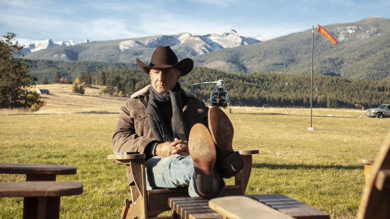 Kevin Costner as John Dutton sitting with his legs crossed and a helicopter in the background on Yellowstone