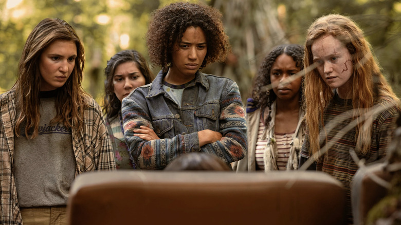 Shauna, Gen, Taissa, Akilah, and Van in the wilderness standing in front of an easy chair on Yellowjackets